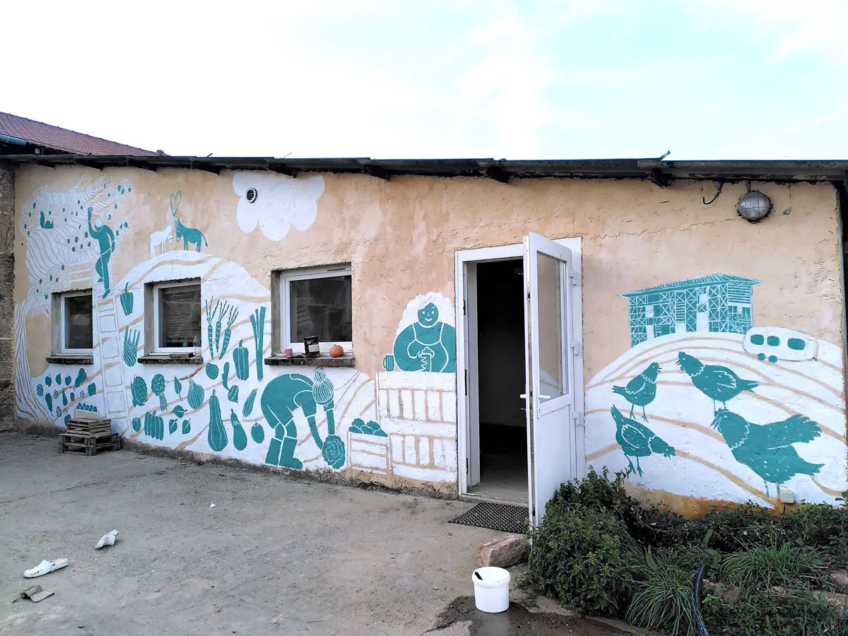 façade en longueur avec trois fenêtres carrées et une porte ouverte. Le mur est peint d'une fresque blanche et bleue avec des légumes, des personnages, un arbre, des poules, une ferme